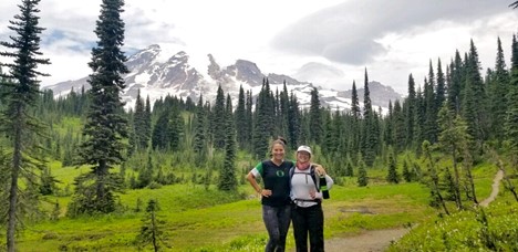 Clara Olson with friend standing in front of Mount Rainier surrounded by evergreen trees.
