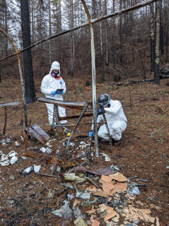 Two people in white coveralls examine a piece of mangled metal after the fires