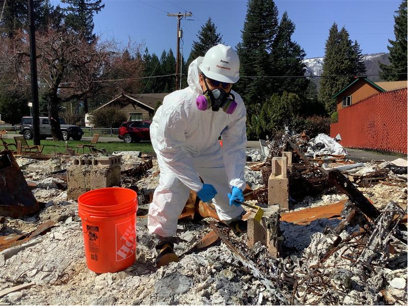 A man wearing a Parametrix hard hat and a white coverall suit works at the site of the fires.