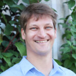 Justin Overdevest wears a blue collared shirt standing in front of greenery