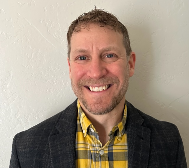 A man smiling wearing a checkered yellow shirt and black jacket