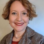 A woman with short hair wearing a red shirt and gray blazer smiling for a headshot photo.