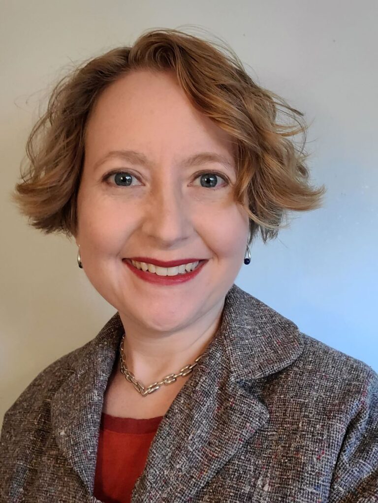 A woman with short hair wearing a red shirt and gray blazer smiling for a headshot photo.