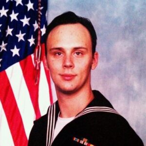 A military headshot of a man wearing a uniform with an American flag in the background.