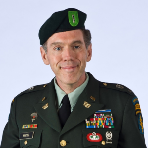 A military headshot of a man wearing a military uniform and hat in front of a bright blue background