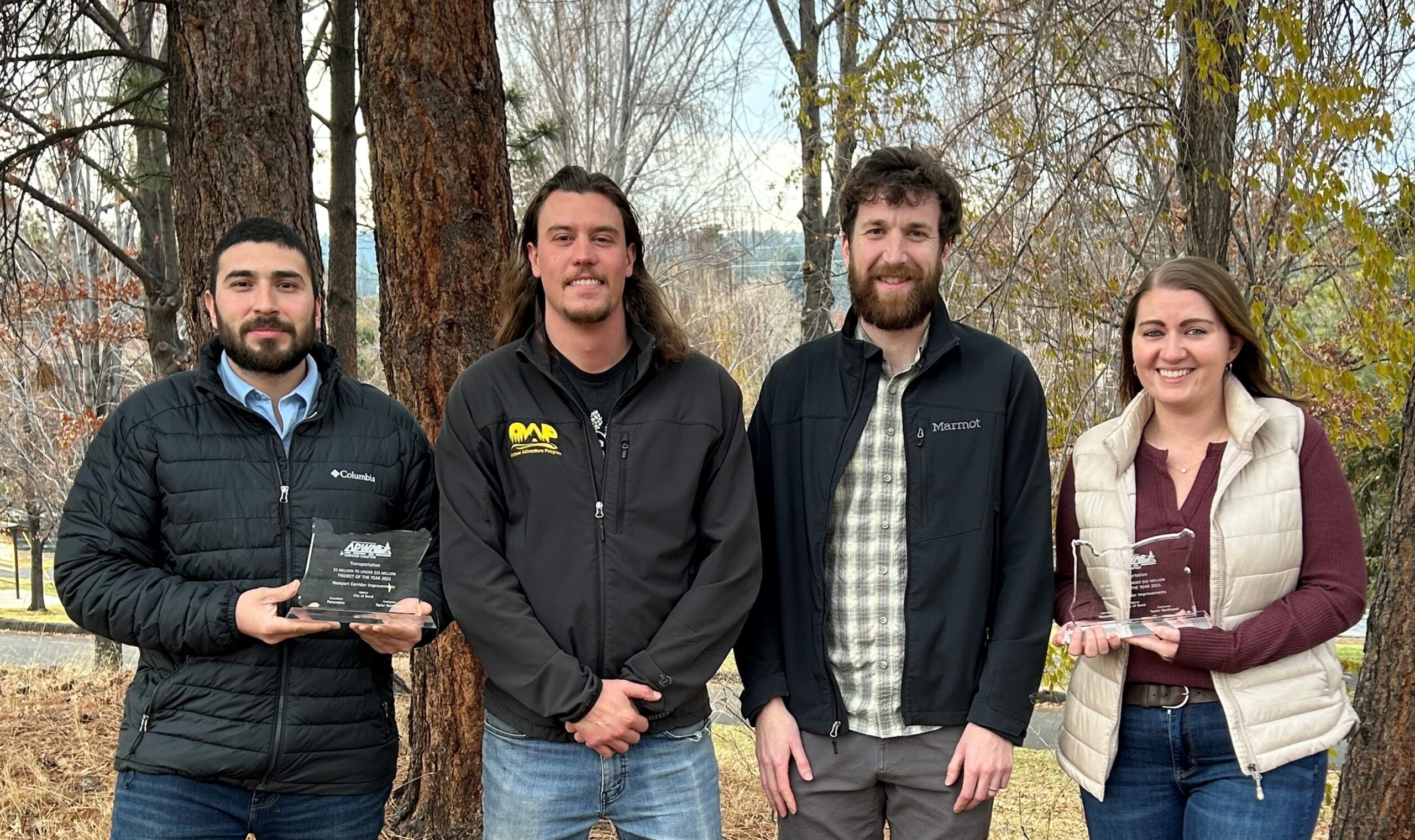 Four people stand outside in front of a treed area posing for a photo, two hold glass trophies.