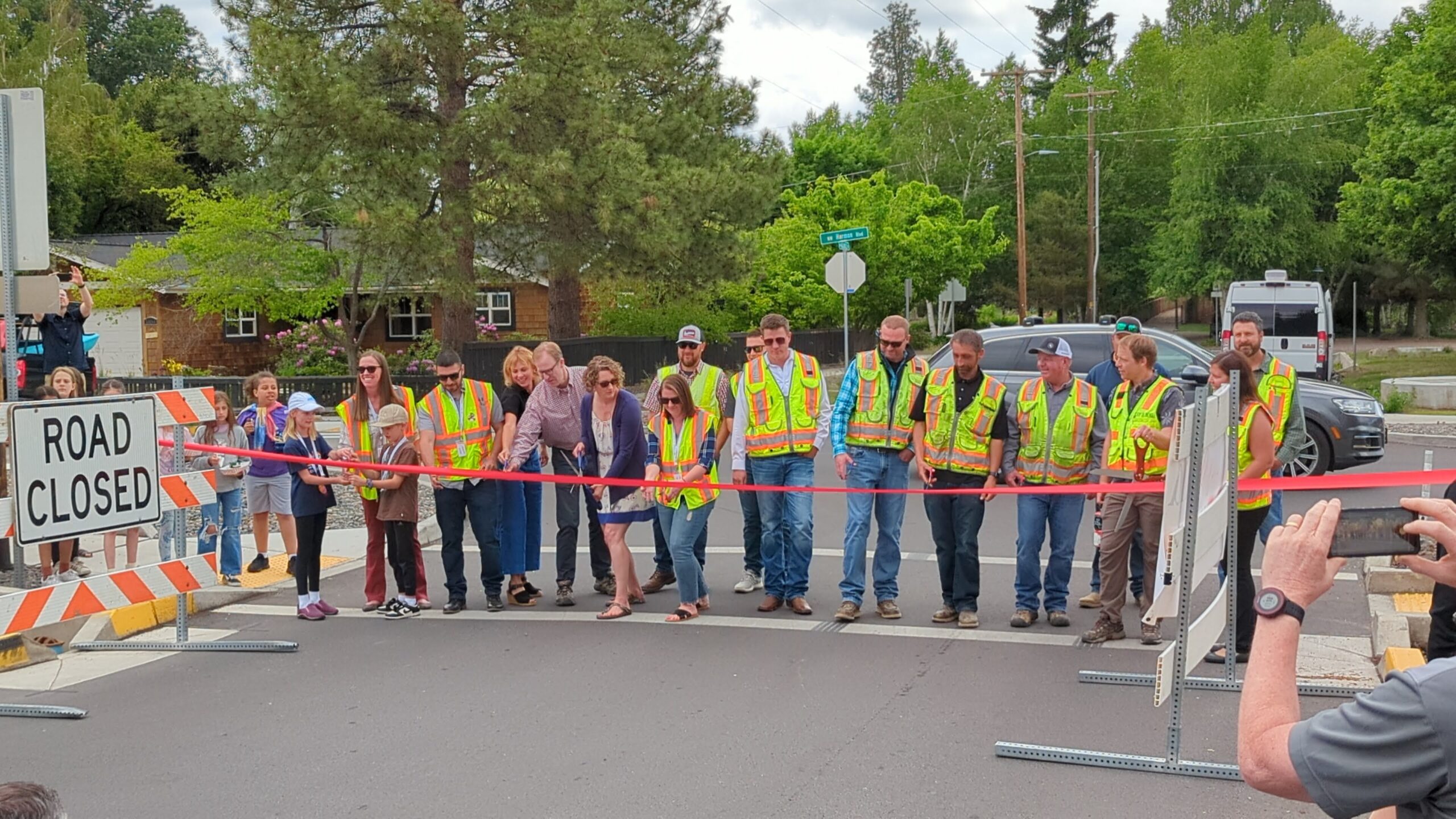 A group of people wearing safety vests stand behind a ribbon. A person in the middle cuts the ribbon.