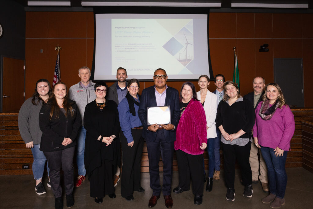A group of people pose for a photo in front of a screen. The person in the middle holds a certificate
