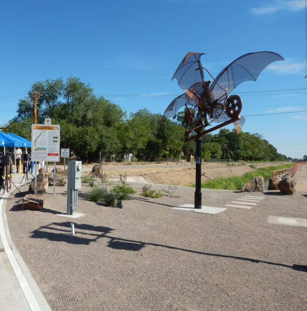 A paved trail with signage and an art feature resembling two bats on a bicycle.
