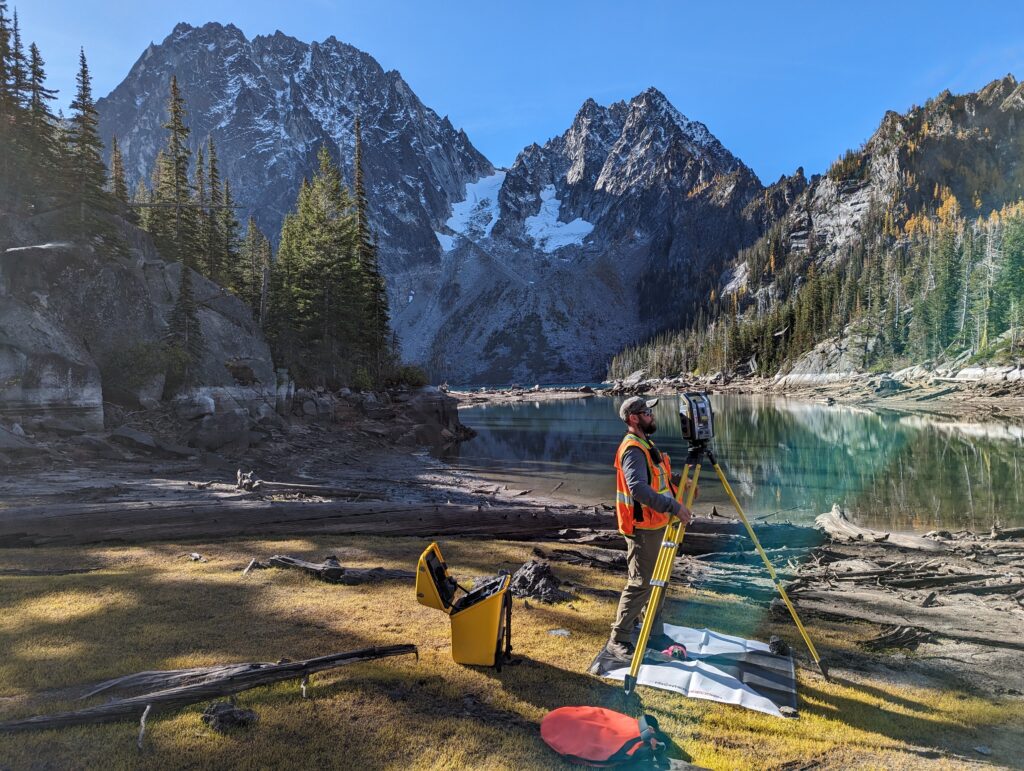 A man works survey equipment in front of a blue lake with snowy hills in the background.
