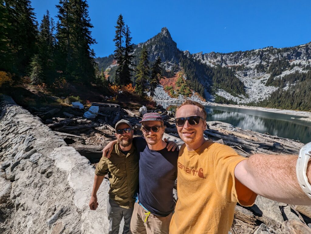 A selfie of three men with a lake and hills in the background.