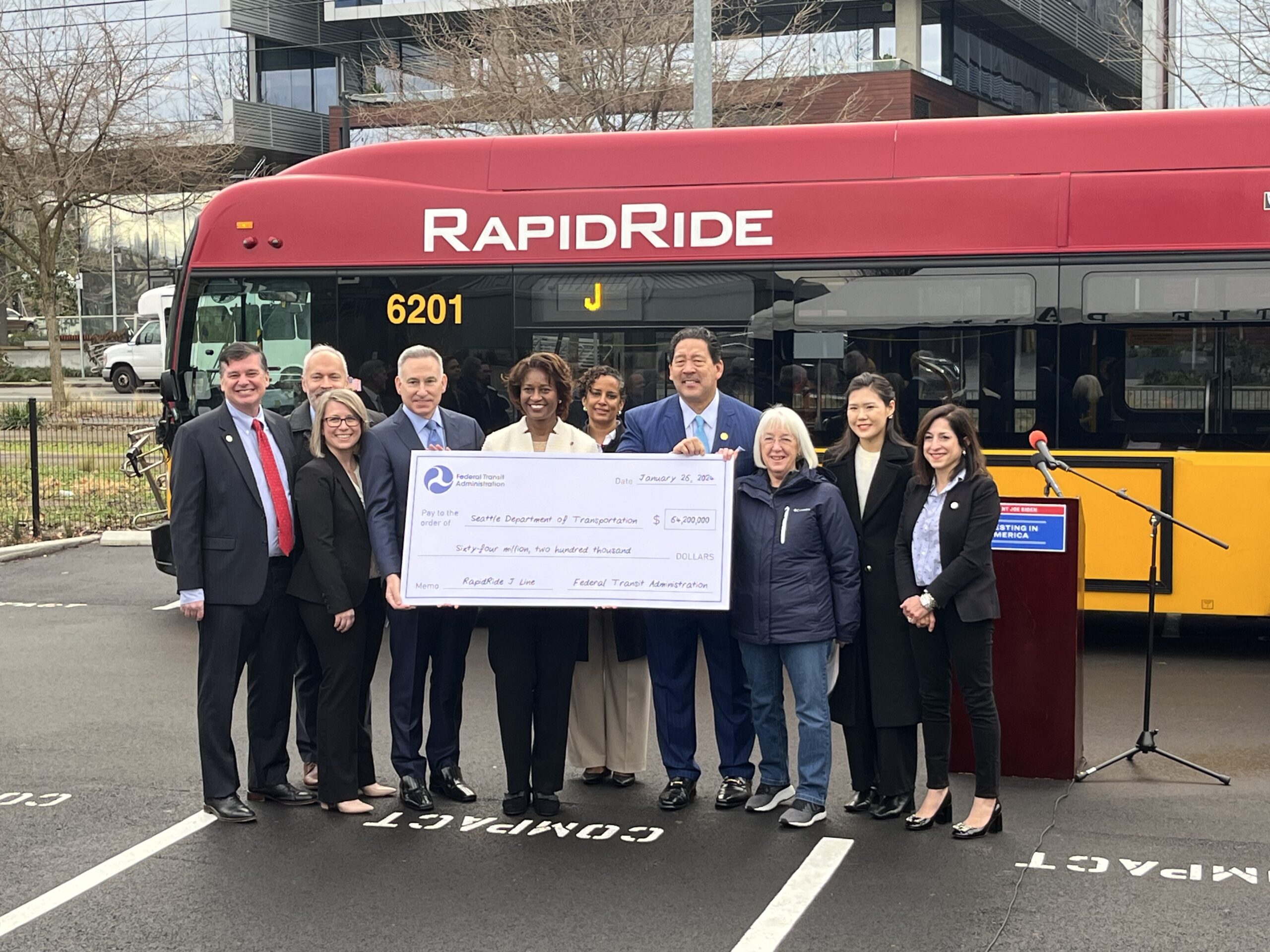 A group of people stand in front of a bus holding a large cardboard check.
