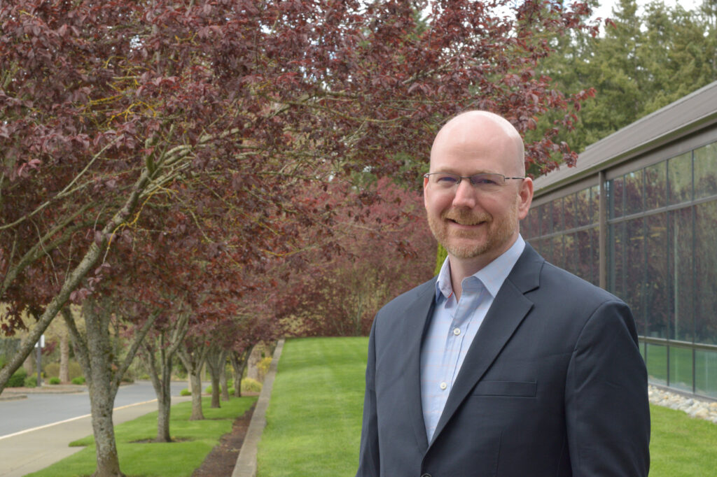 A man wearing a blue collared shirt and suit jacket standing in front of a building and trees.