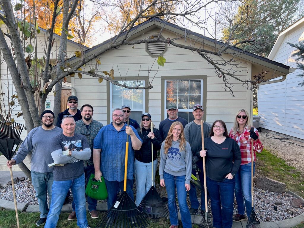 A group of 12 people stand in front of a house holding rakes.