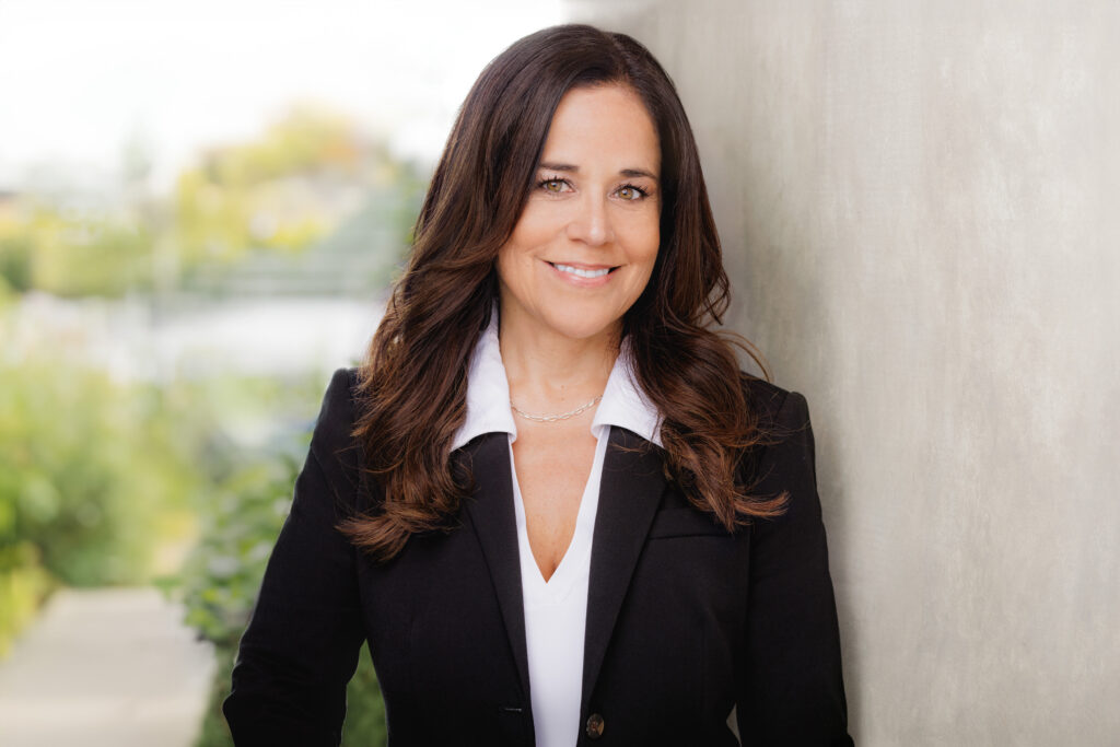 A woman wearing a white collared shirt and black blazer stands smiling in front of a blurred background.