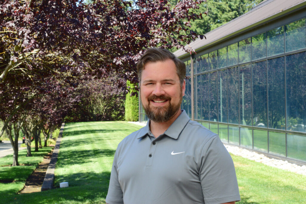 Steven Drangsholt wearing a gray polo standing in front of a grassy outdoor area with trees and a building in the background.