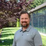 Steven Drangsholt wearing a gray polo standing in front of a grassy outdoor area with trees and a building in the background.