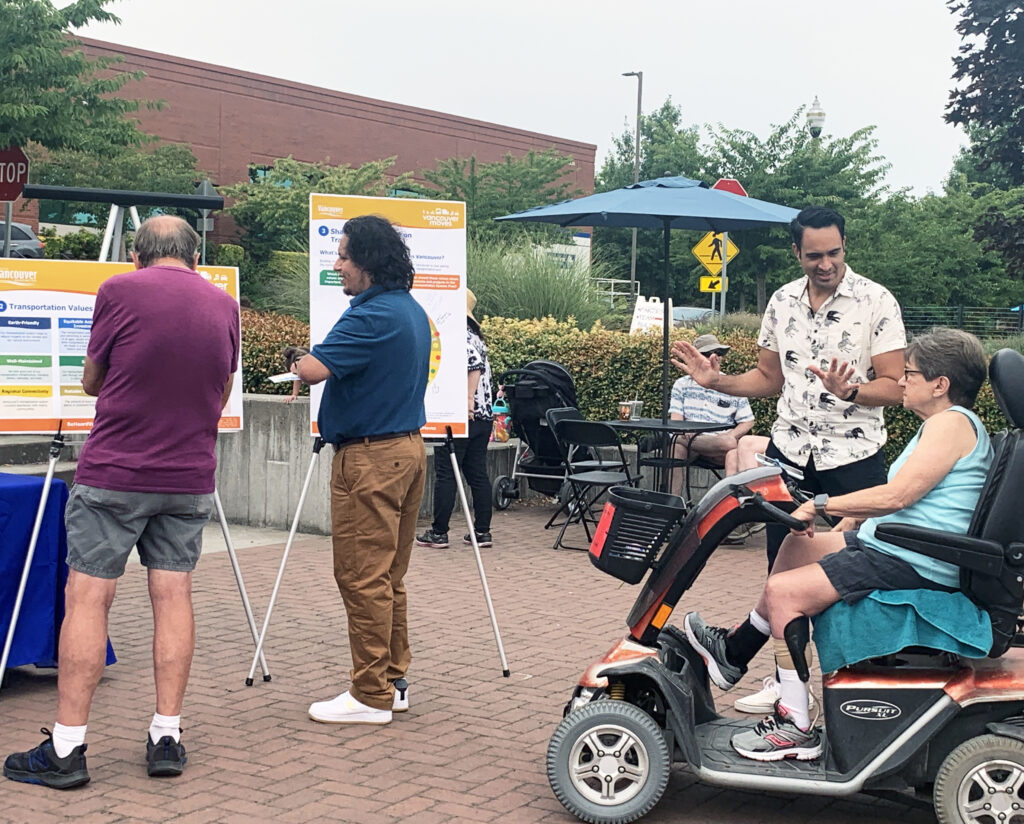 A person standing talking to another person in a motorized scooter. Two other people look at a poster board.
