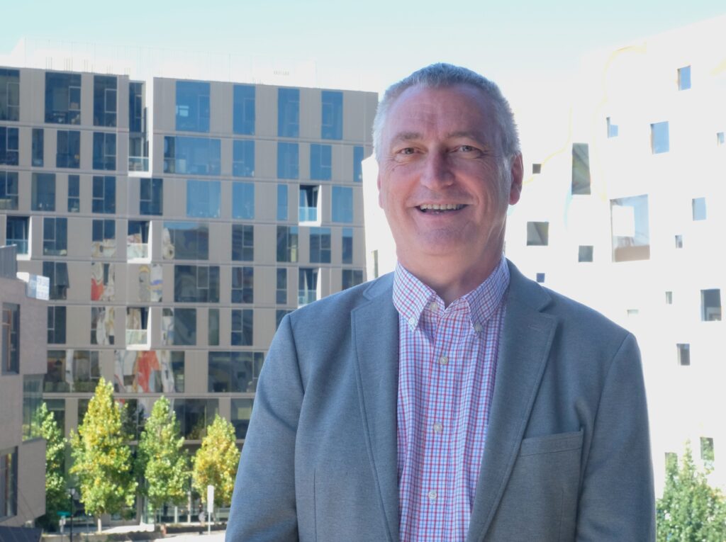 A man poses for a photo in front of a window with buildings in the background.