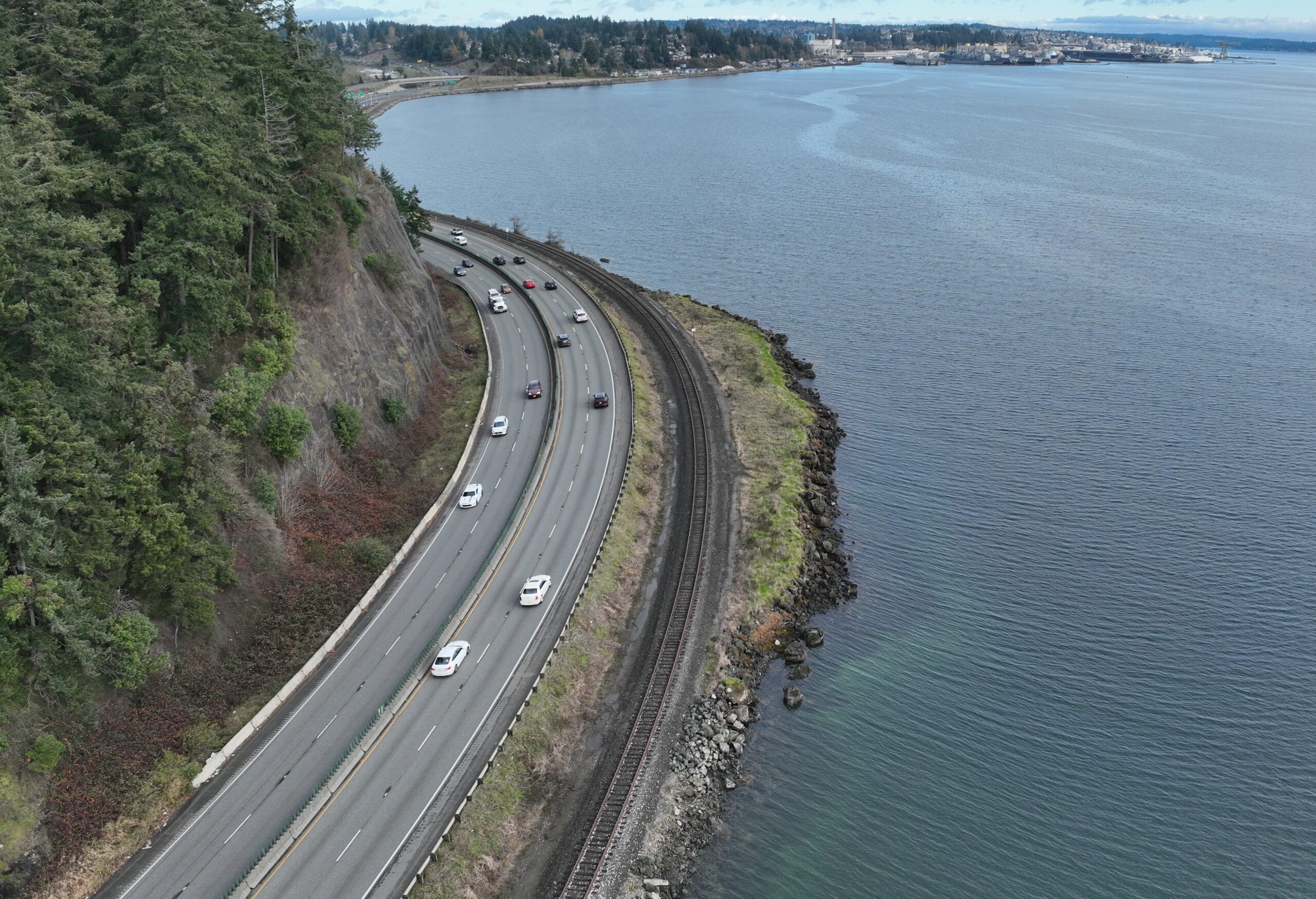 A bird's eye view of a highway that curves between a cliff and a body of water with railroad tracks running parallel to the right.