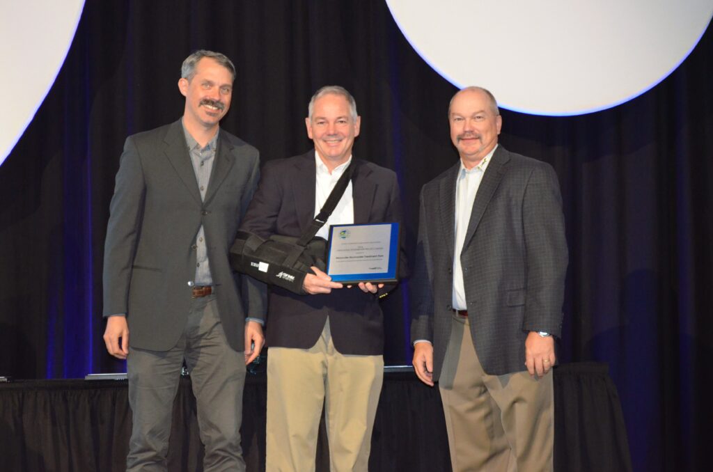 Three people stand on a stage at an award ceremony with the person in the middle holding a plaque.