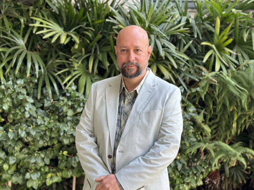 A man wearing a jacket and button up shirt poses in front of green tropical foliage.