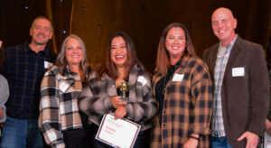 Five people stand on a stage with the person in the middle holding an award certificate