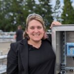 Smiling woman in a black jacket beside an outdoor stainless steel control panel with blue digital meters and cables.