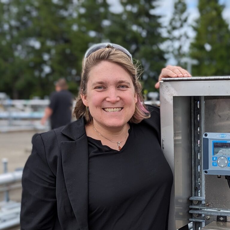 Smiling woman in a black jacket beside an outdoor stainless steel control panel with blue digital meters and cables.
