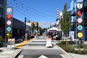 Outdoor street festival with string lights, vendor tents, and people walking between colorful letter-sign columns in a shopping district.