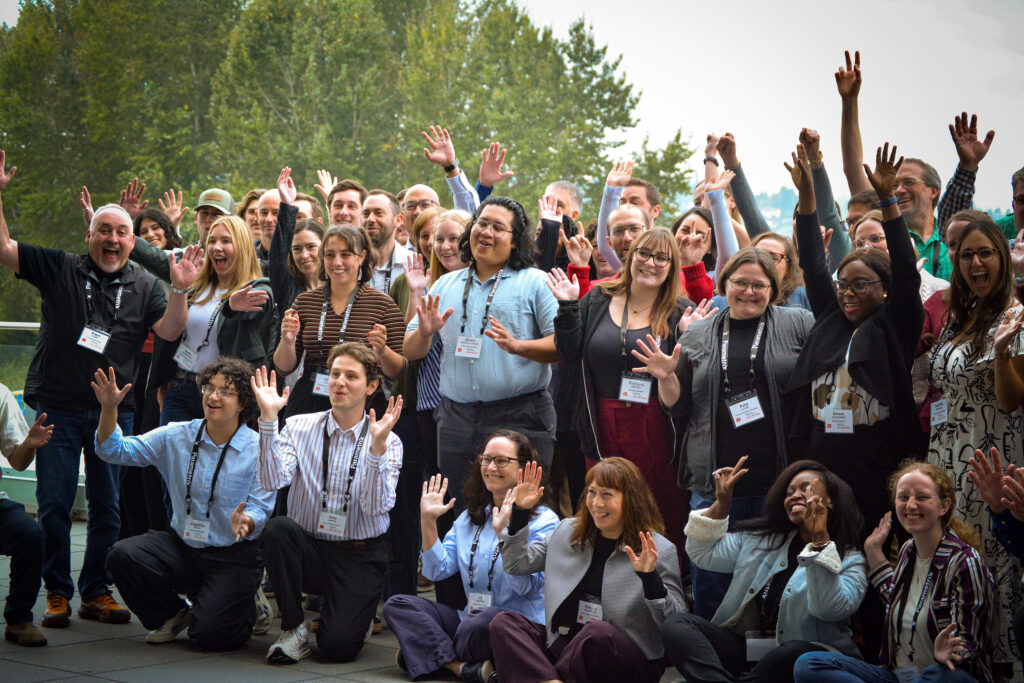 Large diverse group outdoors, many wearing name badges, raising hands and smiling at the camera.