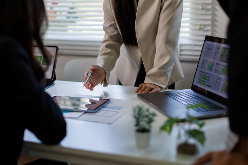 Person in a beige blazer signs a document at a conference table while laptops display charts in the background.