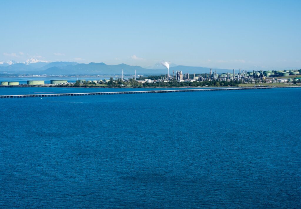 Industrial waterfront with refineries and storage tanks along a blue sea, distant mountains in the background