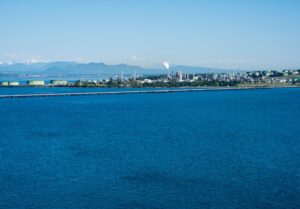 Industrial waterfront with refineries and storage tanks along a blue sea, distant mountains in the background