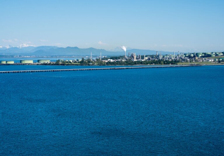 Industrial waterfront with refineries and storage tanks along a blue sea, distant mountains in the background